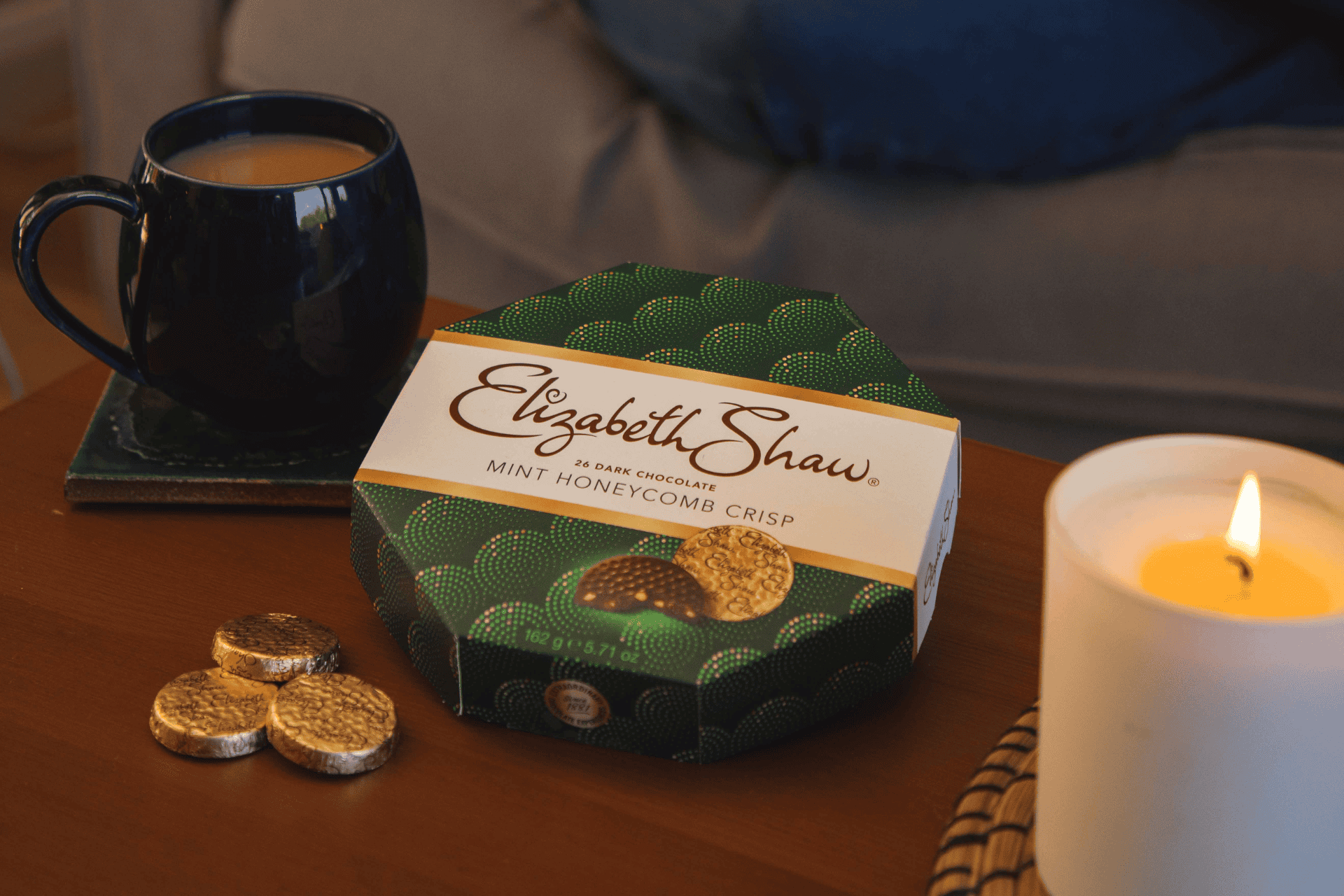 A box of Elizabeth Shaw Dark Chocolate Mint Crisp on a coffee table. Next to it is a cup of tea, a few loose chocolates in gold foil, and a lit candle. In the background you can see the legs of a person sitting on a sofa.