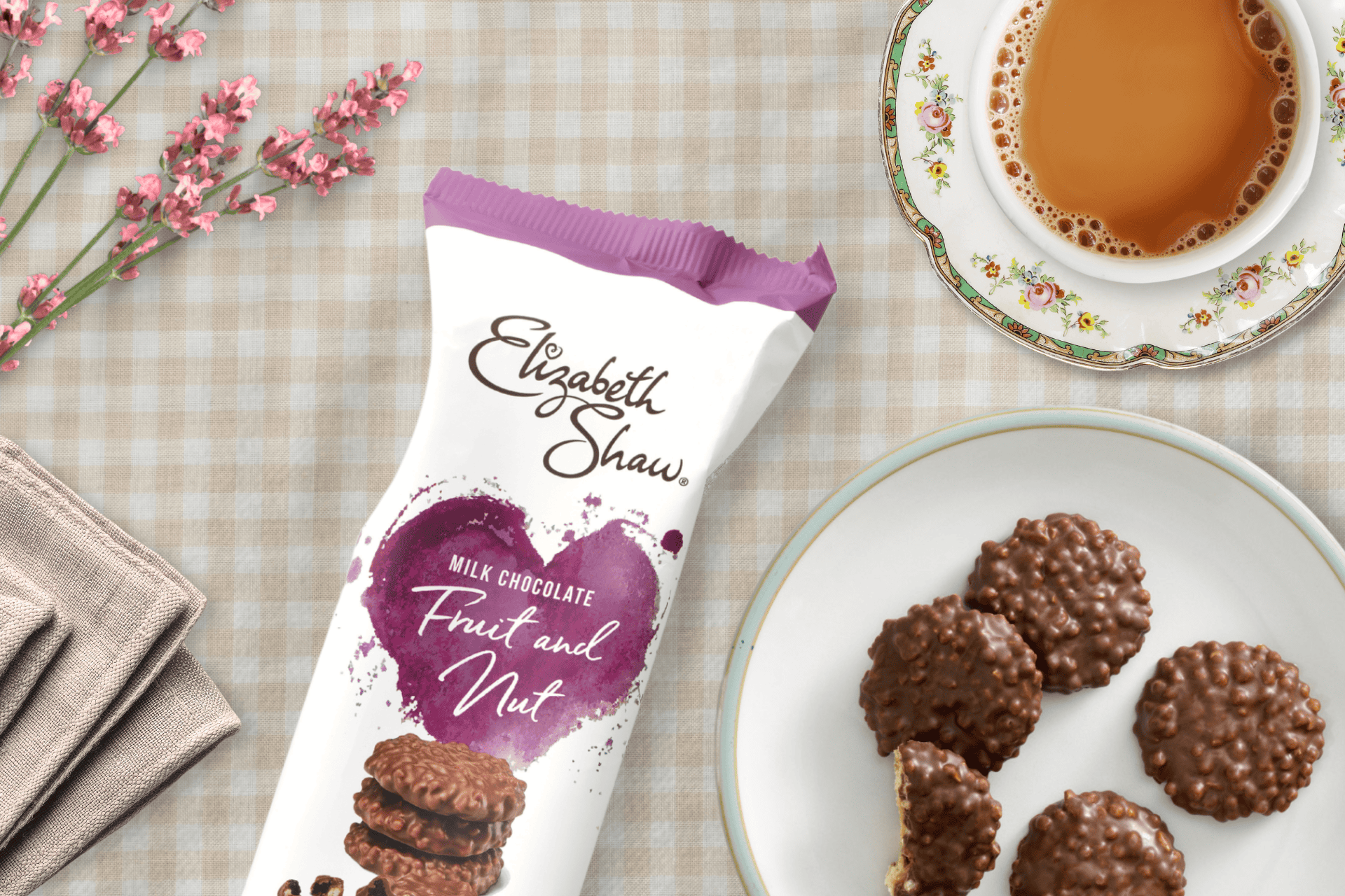 A pack of Elizabeth Shaw Fruit & Nut Milk Chocolate biscuits on a table. There is a beige gingham tablecloth underneath them and a plate of biscuits, a cup of tea, and a bunch of pink flowers alongside them.